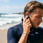 Surfer inserting waterproof ear plugs on a sunny beach.
