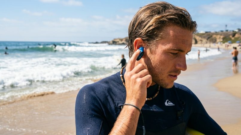 Surfer inserting waterproof ear plugs on a sunny beach.